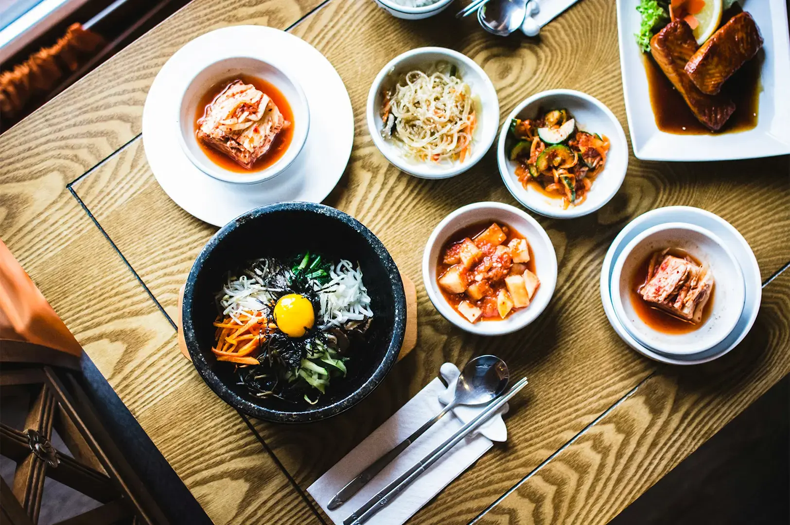 Top-down view of Korean dishes in black and white bowls arranged on top of a wooden textured table beside a window.