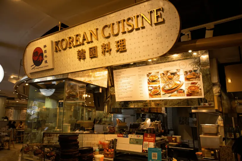 The storefront of the Rui Feng Korean Cuisine stall at ION Orchard's Food Opera food court, featuring a glowing gold sign, glass ordering counter, and an illuminated menu board showcasing hotplate and soup set meals.