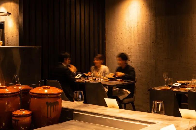 An atmospheric, candid shot capturing the dining ambiance at Kemuri. In the sharply focused foreground, traditional glossy brown ceramic pots rest on the concrete counter. In the softly blurred background, three diners enjoy a meal at a dark table against a textured grey wall, illuminated by a warm wall sconce.