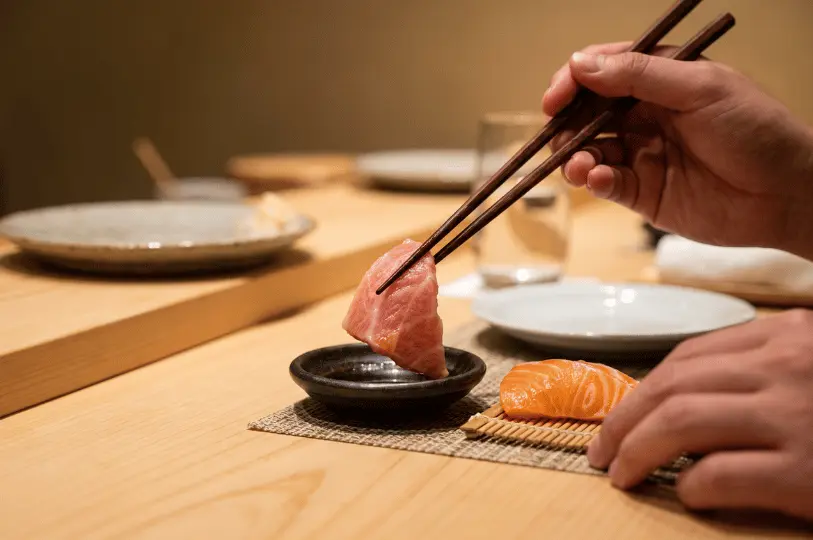 A close-up shot of a diner's hands using wooden chopsticks to delicately dip a piece of marbled fatty tuna sashimi into a small, dark dish of soy sauce. A piece of salmon sashimi rests on a mini bamboo mat nearby.
