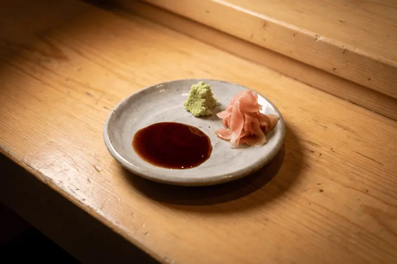 A minimalist, rustic ceramic condiment plate resting on a wooden surface, neatly presenting a pool of dark soy sauce, a small mound of freshly grated green wasabi, and a pile of pickled pink ginger.