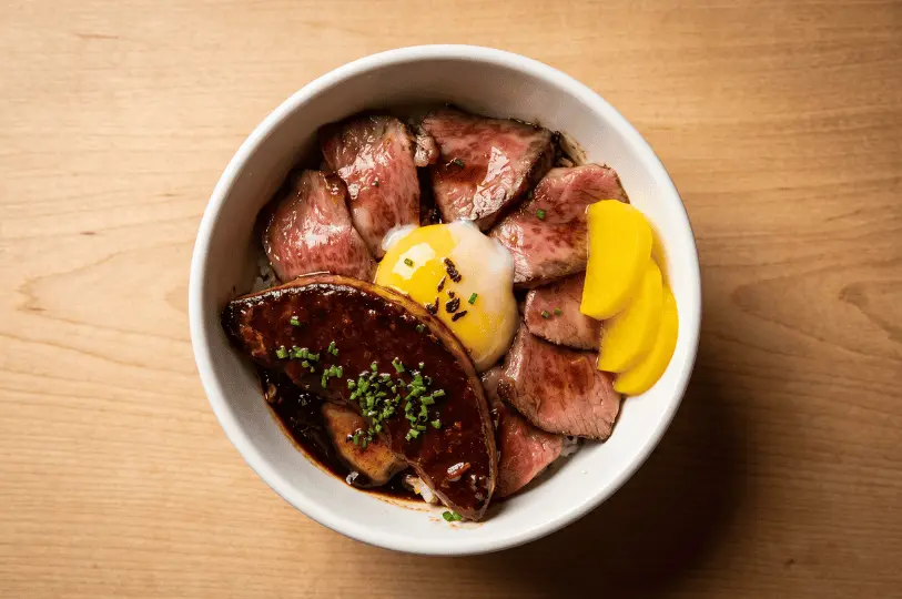 Top-down view of a rich rice bowl covered in seared sliced beef, a large piece of dark, glazed foie gras, a soft-boiled egg, and bright yellow pickled radish slices.