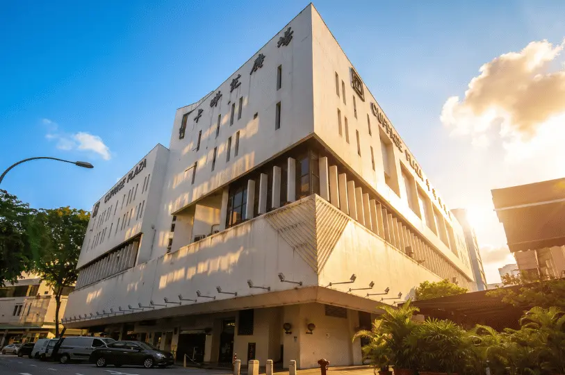 A low-angle, exterior shot of the Cuppage Plaza building, a multi-story white commercial complex with unique geometric architecture, bathed in the warm, golden light of the sun.