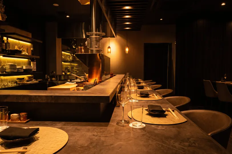An intimate, low-angle perspective looking down Kemuri's polished concrete dining counter. The scene is bathed in warm, low-key lighting from hanging pendant lamps and the glowing hearth behind the glass. Empty wine glasses, woven placemats, and dark napkins are meticulously arranged, emphasizing the quiet, upscale ambiance.