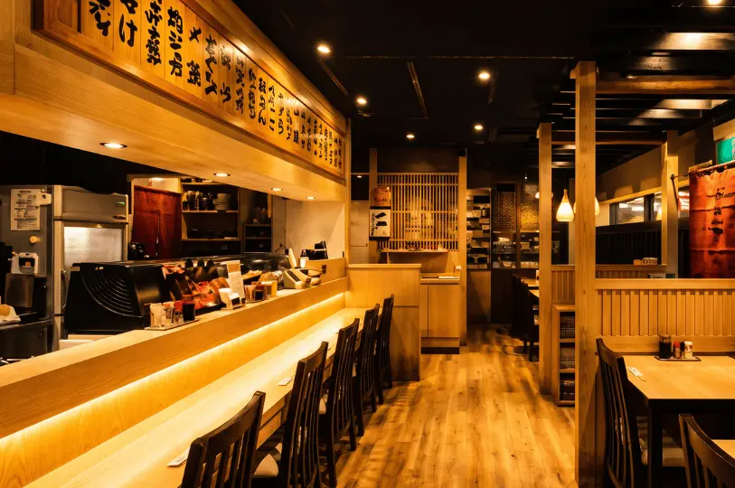 The warm, inviting interior of Shinjuku Restaurant, showcasing a classic izakaya layout with a long, illuminated wooden sushi counter lined with dark chairs on the left, and cozy, traditional wooden booth seating on the right.