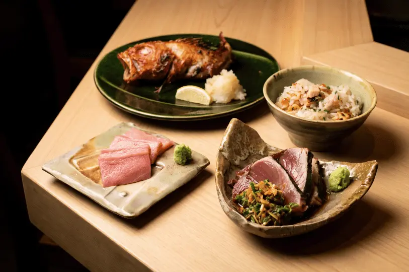 An elegant flat-lay composition of four Japanese dishes on a light wood surface. The frame includes a whole crispy fried fish with grated daikon on a dark green plate, a bowl of mixed rice studded with salmon and ikura, a square ceramic dish of vibrant pink fatty tuna sashimi, and a rustic bowl presenting seared bonito slices topped with fresh garnishes.
