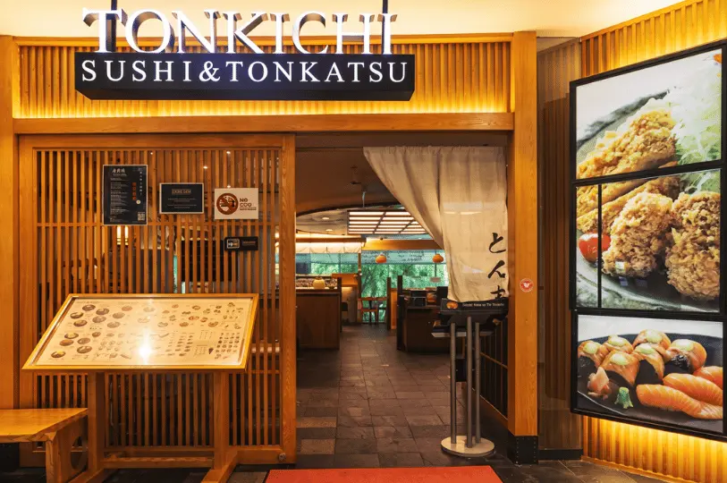 The entrance to the Tonkichi Sushi & Tonkatsu restaurant, featuring a traditional wooden slatted facade, a white noren curtain over the doorway, and large illuminated menu photos on the right wall.