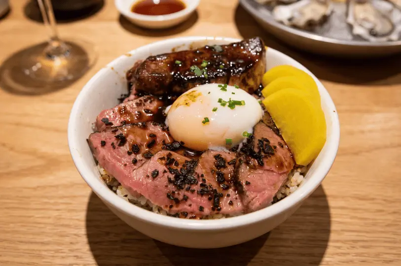 A close-up side angle of a savory rice bowl topped with seared sliced beef, a thick piece of glazed pan-seared foie gras, a soft-boiled onsen egg, and slices of yellow pickled radish.