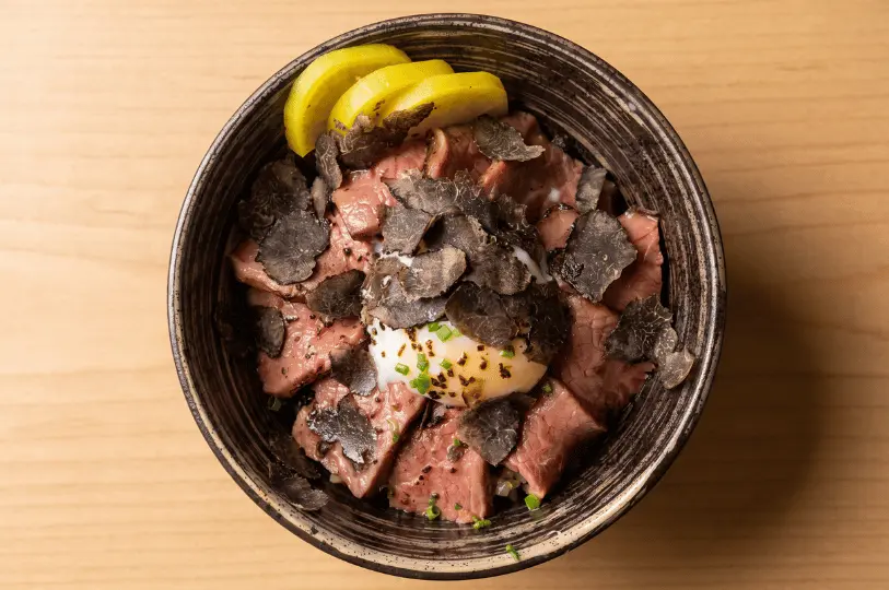 Top-down view of a decadent beef donburi (rice bowl) heavily layered with shaved black truffle over sliced beef, featuring a soft-boiled egg in the center and yellow pickled radish on the side.