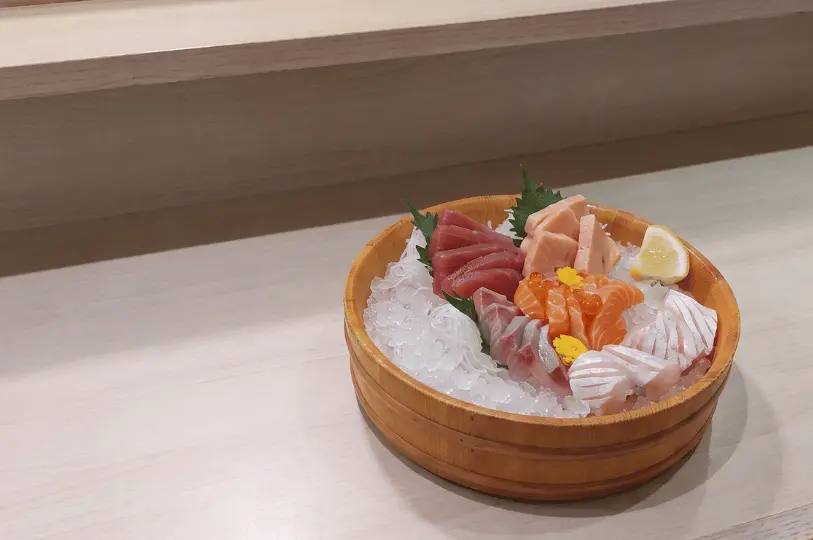 A round wooden bowl filled with crushed ice, displaying a vibrant and fresh assortment of sashimi. The selection includes bright red tuna, orange salmon topped with roe, and intricately scored white fish, elegantly garnished with green shiso leaves, tiny yellow flowers, and a lemon wedge.