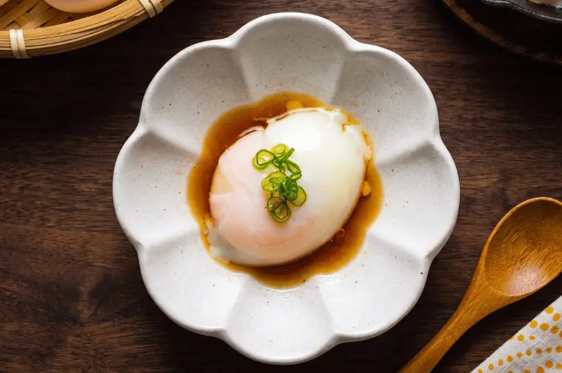A soft-boiled onsen egg resting in a savory soy-based broth, garnished with chopped green onions, and served in a delicate, flower-shaped ceramic dish alongside a wooden spoon.