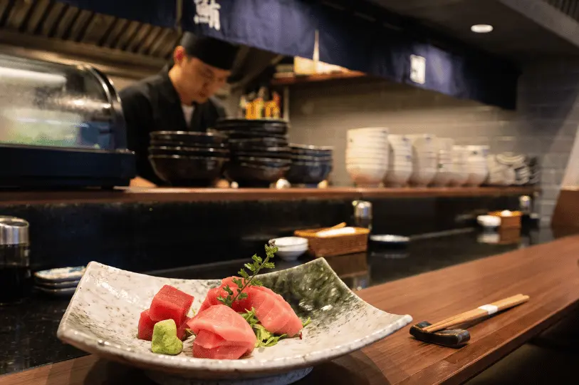 Thickly sliced bright red and pink tuna sashimi served on a curved, rustic ceramic dish with a side of fresh wasabi. The plate sits on a dark wooden sushi bar, while a chef in a black uniform is blurred in the background surrounded by stacks of bowls.