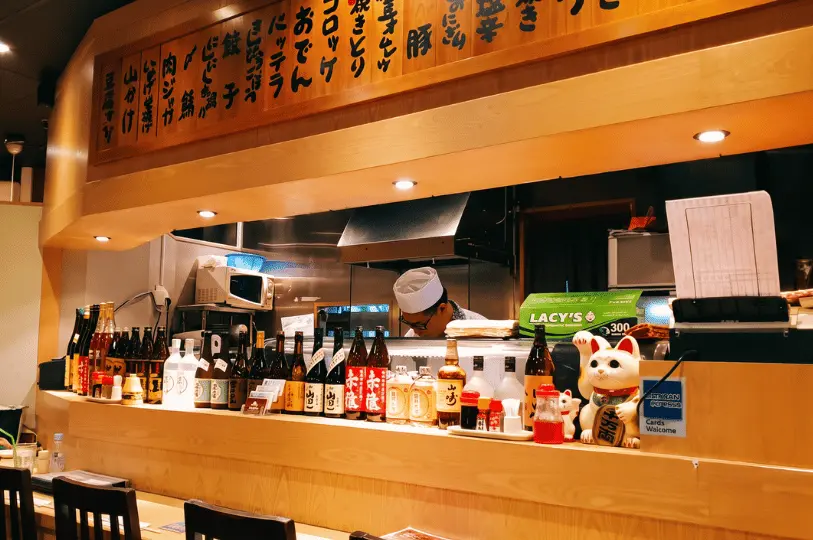 A close-up view of the wooden dining counter at Shinjuku Restaurant, cluttered authentically with sake bottles, condiments, and a Maneki-neko lucky cat figurine. A chef in a white uniform works in the kitchen behind the counter, beneath a wooden board displaying handwritten Japanese menu items.