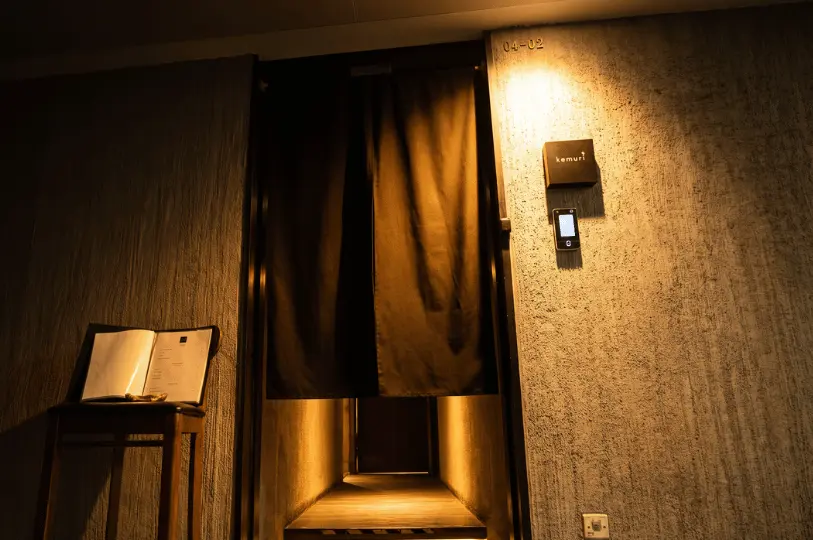 A dimly lit exterior shot of the Kemuri entrance, featuring rough, raw concrete walls illuminated by a single warm spotlight. A dark, split noren curtain conceals a glowing, narrow wooden corridor leading inside. To the left, a wooden stand holds an open menu, while a minimalist black