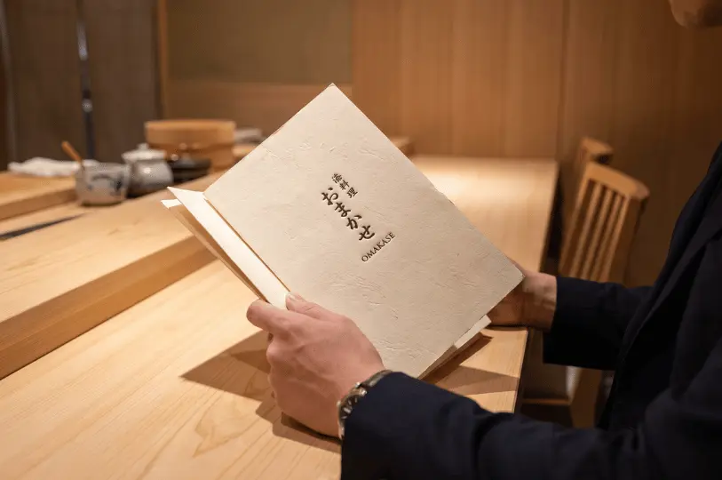 A guest seated at a smooth wooden sushi counter holding open a textured, light-colored menu. The cover of the menu clearly reads 