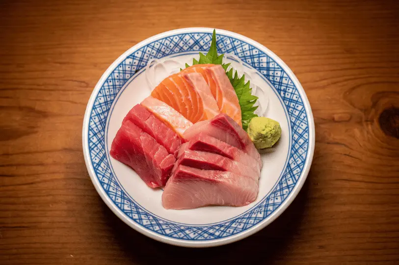 A top-down view of a blue and white patterned bowl containing thick, fresh cuts of raw salmon, tuna, and yellowtail sashimi, garnished with a green shiso leaf and a dollop of wasabi.