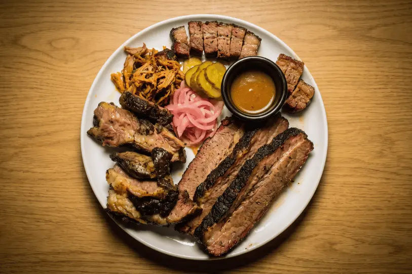 A top-down macro shot highlighting the rich textures of a smoked meat platter on a light oak table. The round white plate is loaded with thick, dark-barked brisket slices, cubed pork belly, pulled pork, and bright pink pickled red onions and pickles, all orbiting a small dark ramekin of yellow mustard sauce.