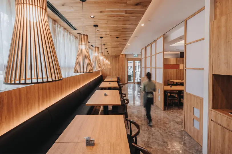 The bright and serene interior of a Japanese dining establishment, featuring light wood paneling, slatted wooden pendant lights over booth seating, and traditional shoji-style sliding doors.