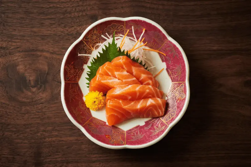 An overhead view of vibrant orange salmon sashimi slices elegantly arranged on a decorative red and white plate with gold floral patterns, garnished with a shiso leaf, shredded daikon radish, and a small yellow flower.