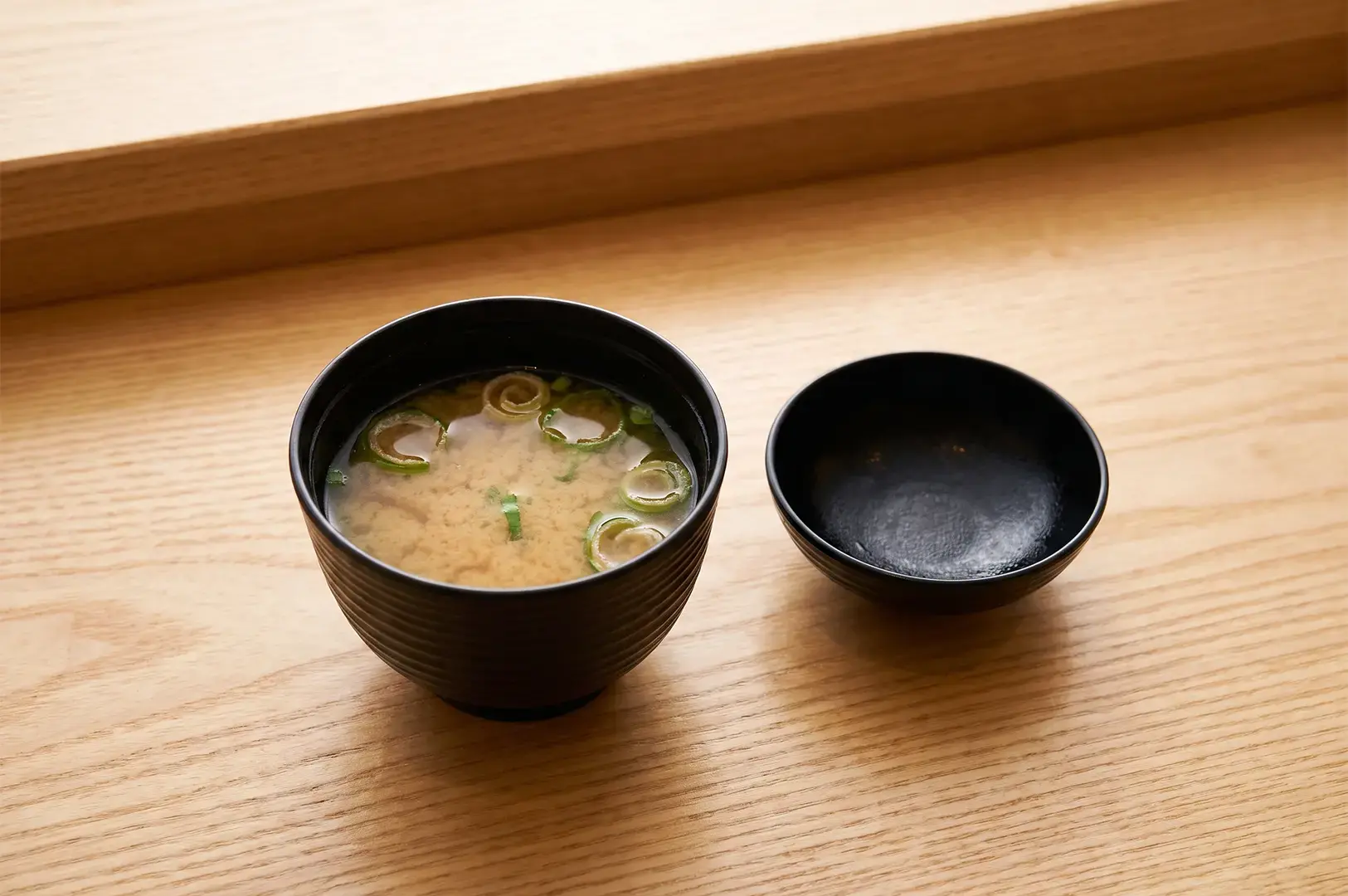 Miso Soup in a black bowl with its cover by the side on a wooden countertop served at Ginza Sushi Ichi.