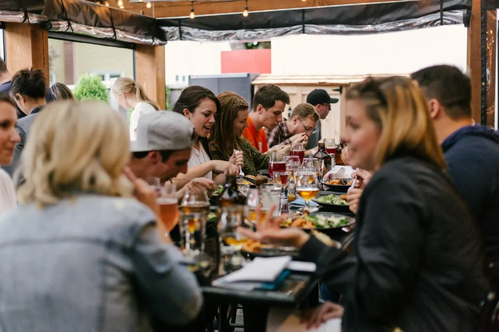 A group of people sat on a long table filled with food ang glasses while eating and conversing in a lighthearted mood inside a public establishment.