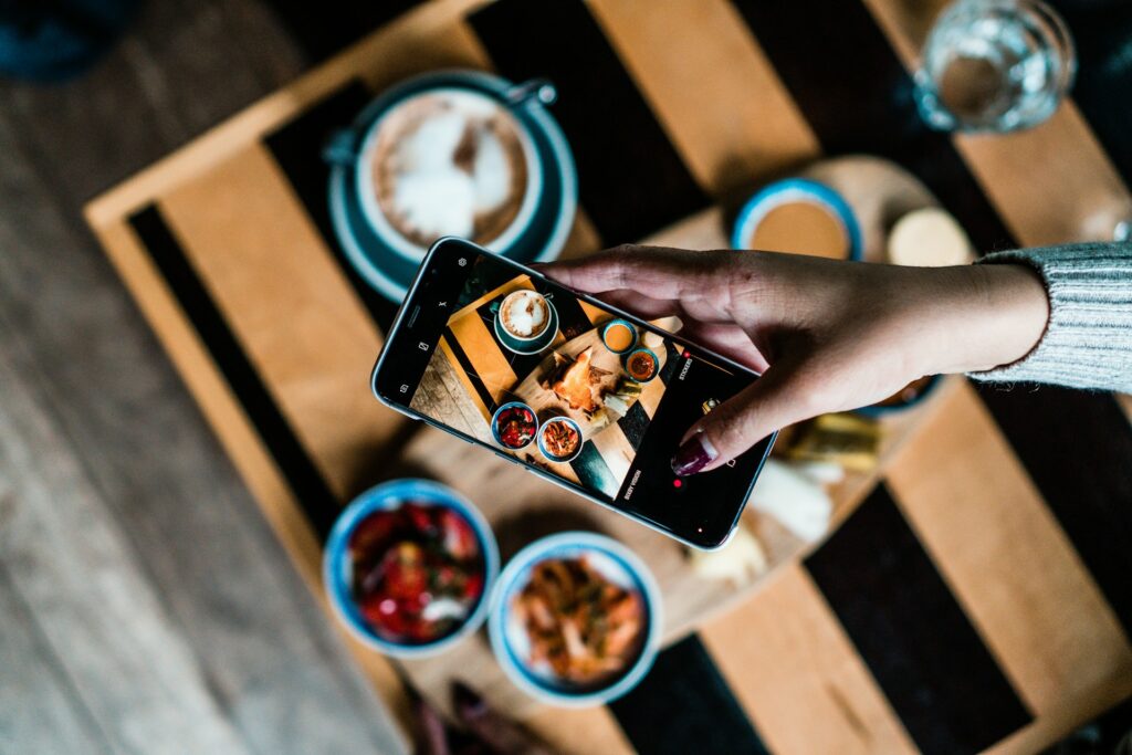 A hand holding a smartphone to capture a photo of a dish placed on top of a wooden table blurred in the background.