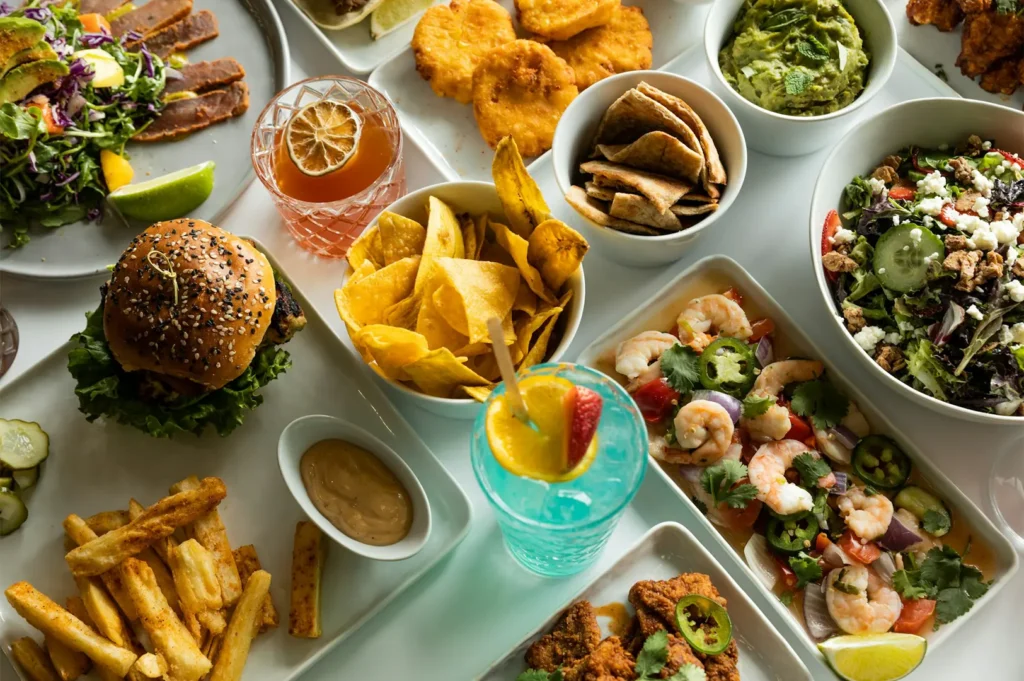 Flat lay wide shot of a variety of food including burgers, nachos, fries, gravy, shrimp, and an assortment of leafy greens all placed on a white table.