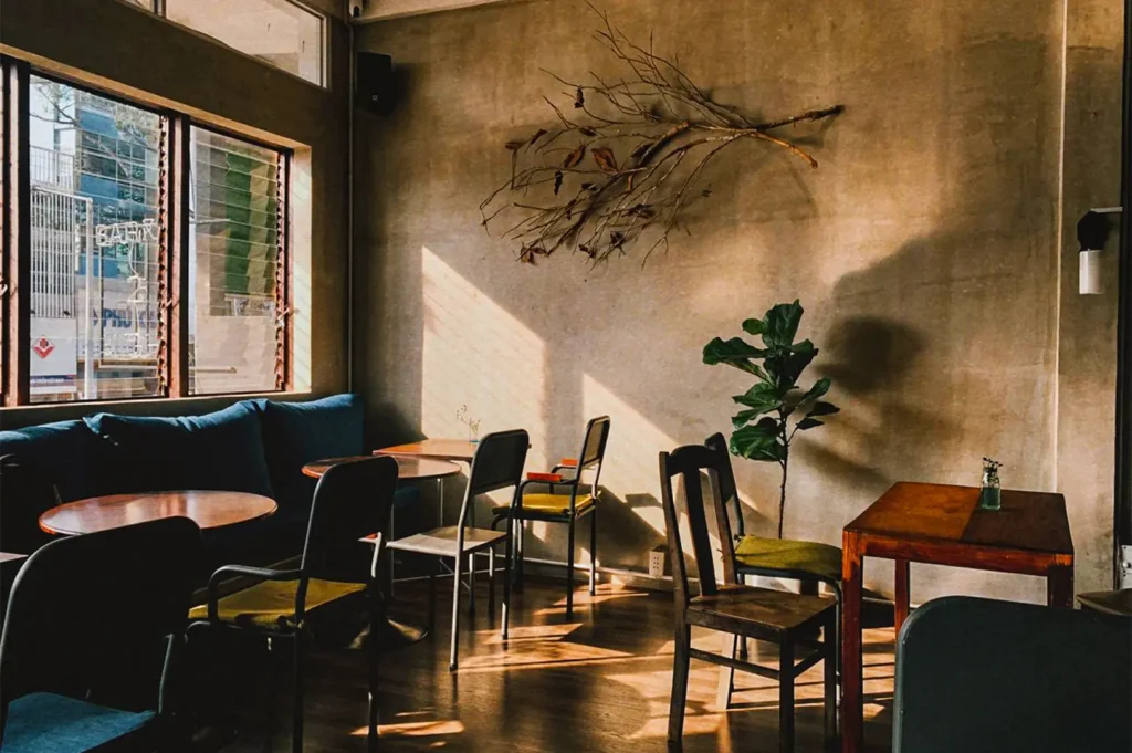 Cozy cafe interior with empty tables, wooden chairs, plants, and warm sunlight streaming through the windows.