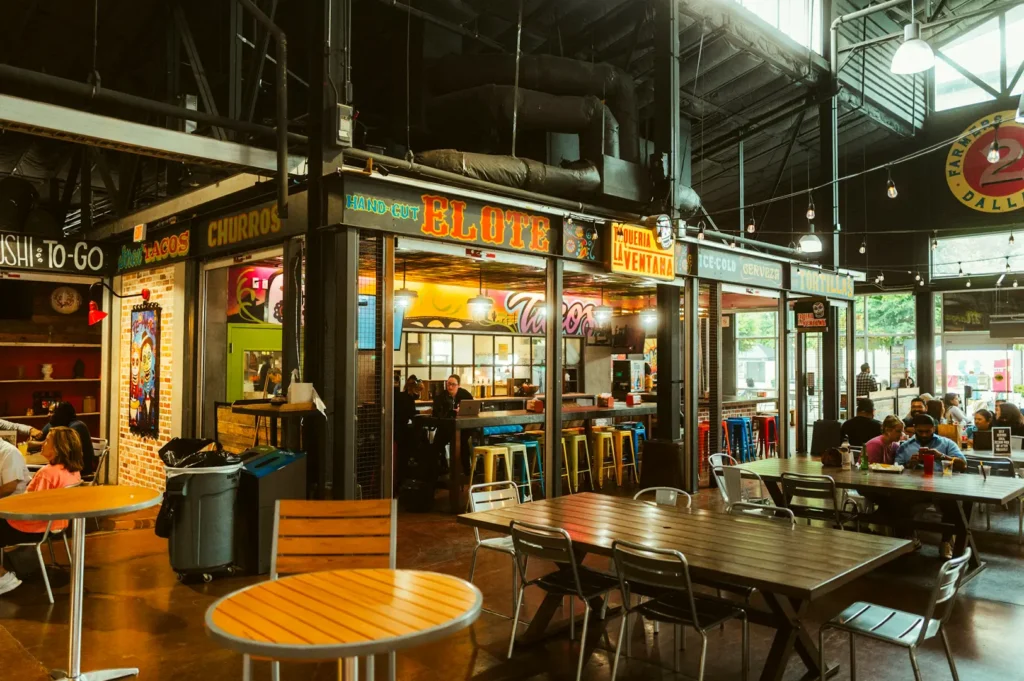Wide angle shot of a busy food court with different food stalls lined up in the background surrounded by customers with empty chairs and tables in the foreground.