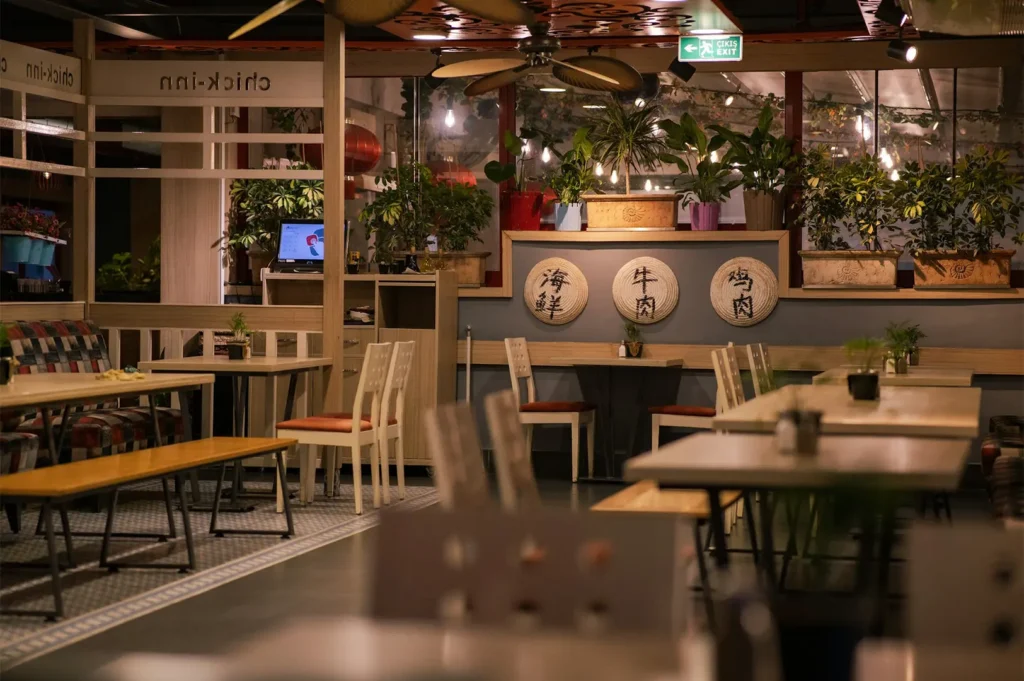 A cozy restaurant interior with empty white chairs and tables decorated with green potted plants and illuminated with warm lighting.