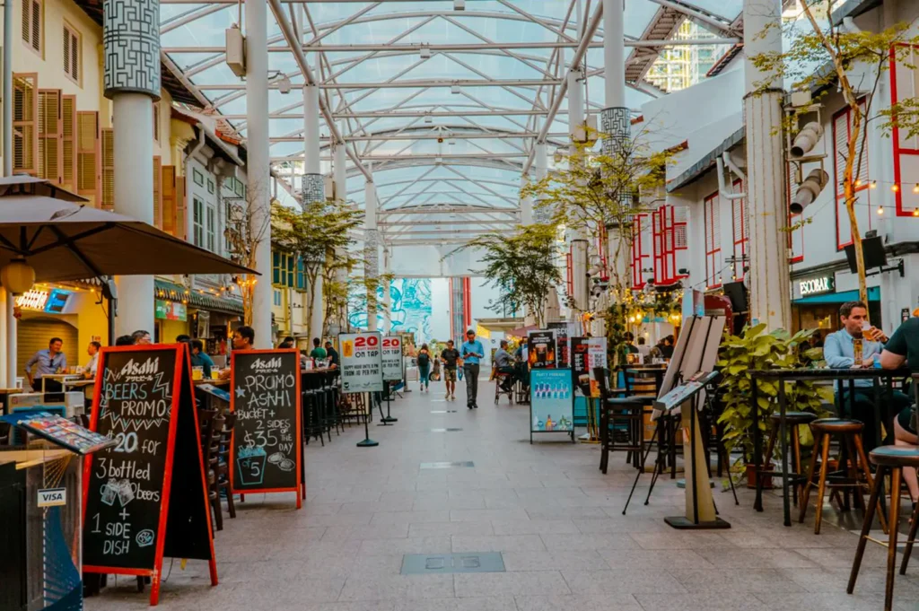 Cafes and restaurants lining two sides under a glass ceiling with busy people talking and dining on tables and chairs.