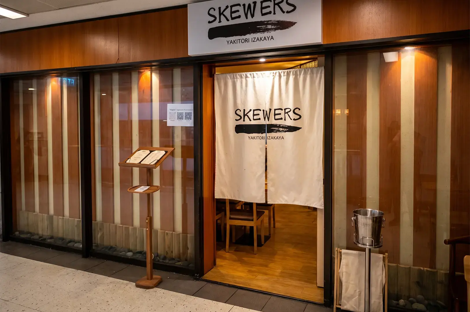 Skewers Yakitori exterior shot with patterned wood panels behind glass and its logo visible on its upper signage and curtain at the entrance.