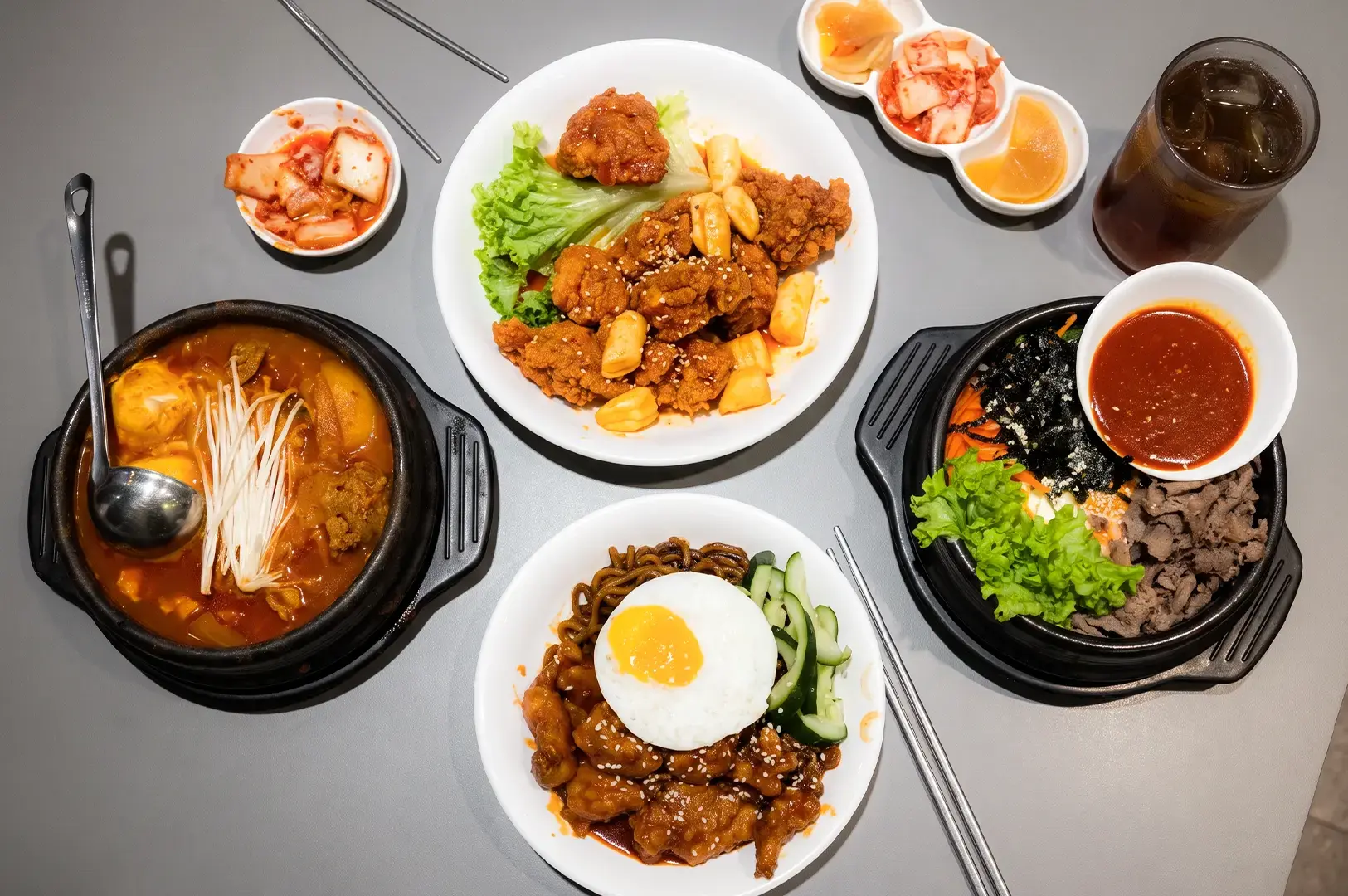 Flat lay shot of Pocha Korean Street Dining menu items including soup pots, chicken, and noodles with side dishes placed on a grey table.