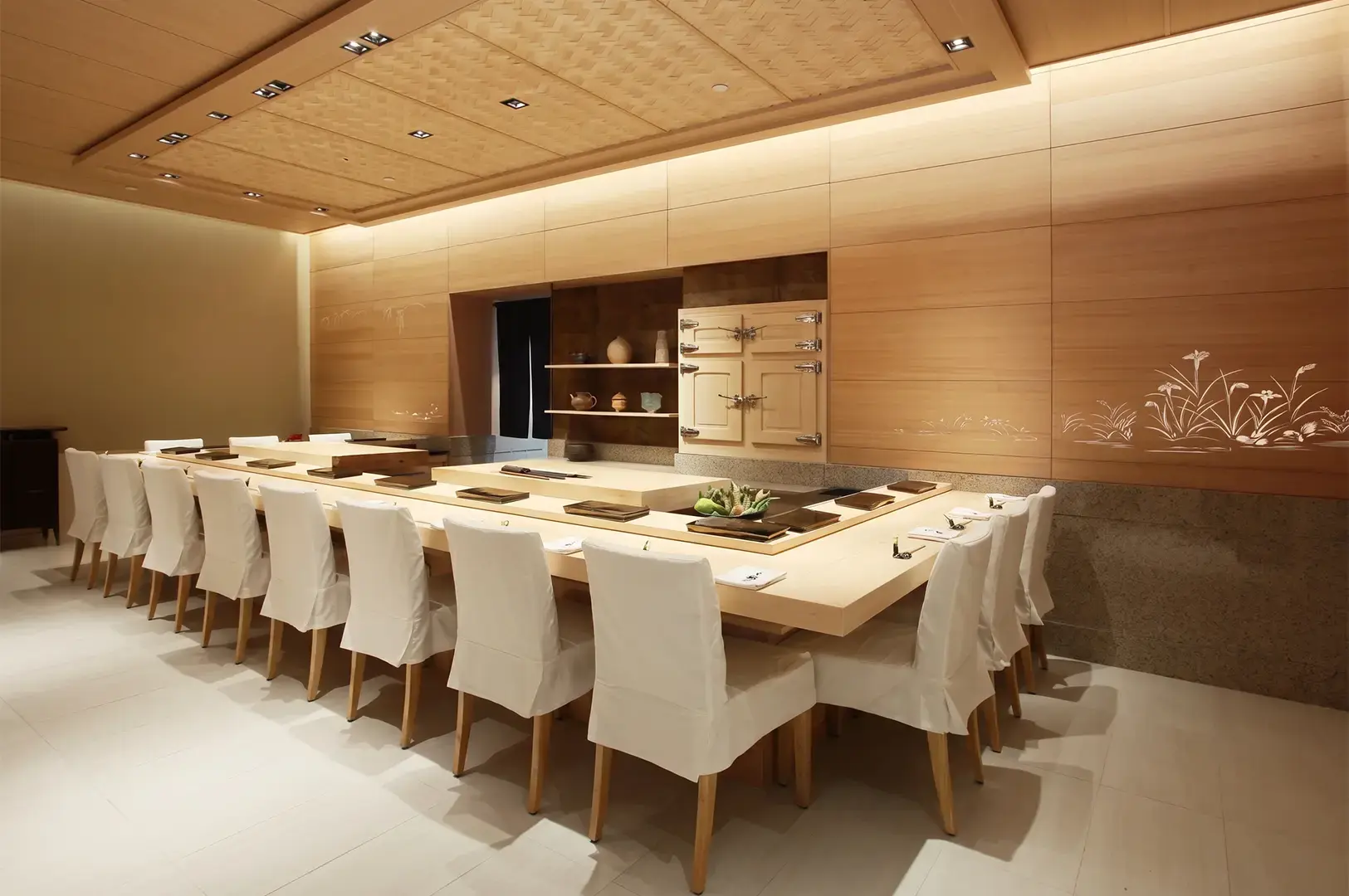 Ginza Sushi Ichi interior with neatly prepared wooden table and white chairs lined with wooden paneled walles and shelves and warm, cozy lighting.