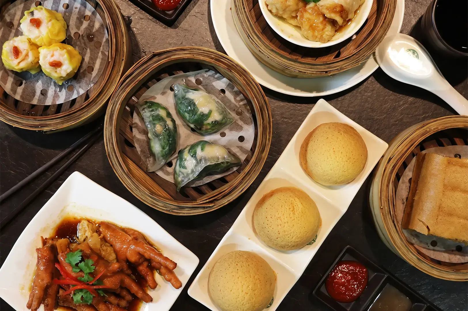 Flat lay high angle shot of Tim Ho Wan's famous dishes including Pork Buns, Dim Sums, and Pork and Shrimp Dumplings with sauces arranged on a dark textured table.