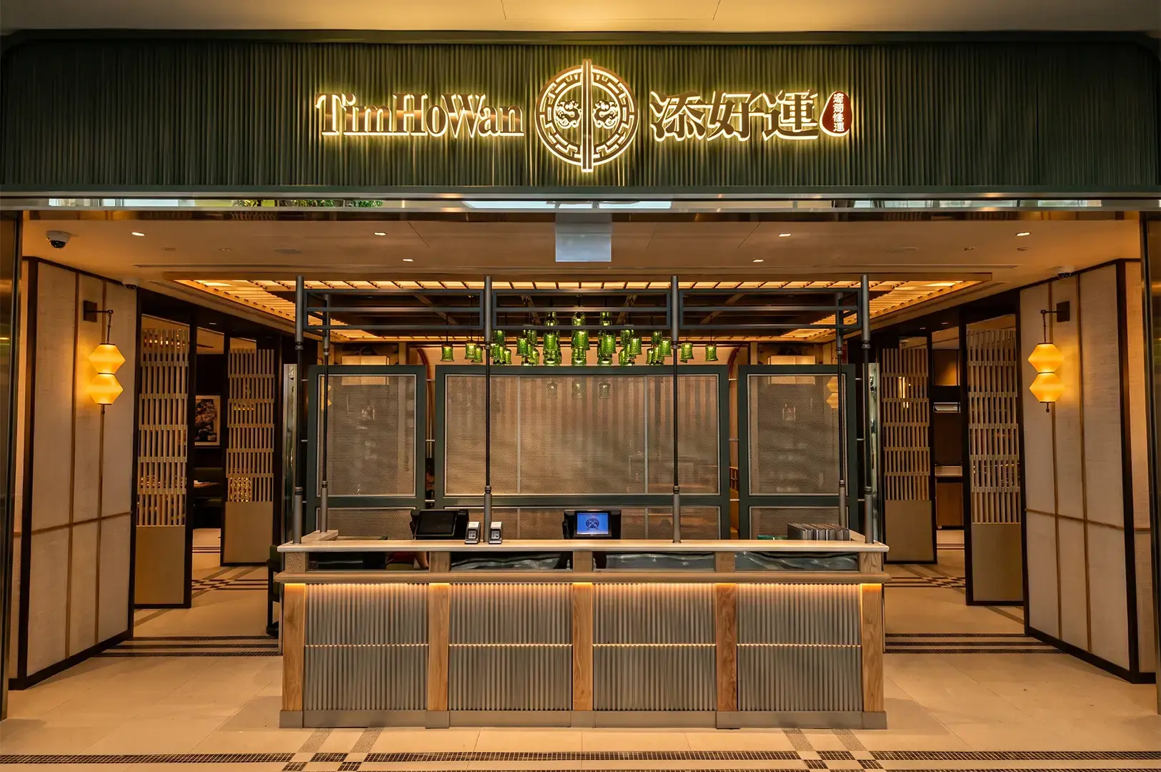 Wide angle shot of Tim Ho Wan exterior, with the counter at the front and entrances on both sides visible, its logo signage and lamps lit by warm yellow lights.