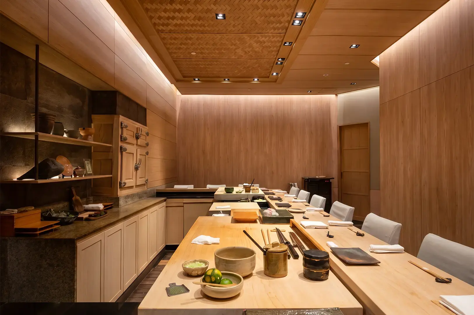 Ginza Sushi Ichi interior showing the counter with prepared kitchenware on top, shelves at the back, and empty chairs in front lit with warm lights.