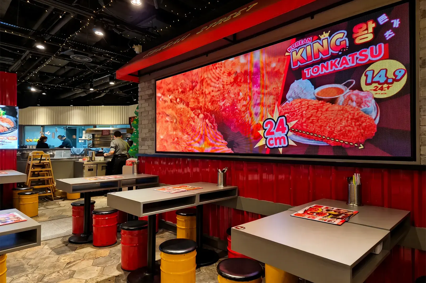 A row of tables and chairs inside Pocha Korean Street Dining with a promotional poster displayed on a side wall covered in red panels and the busy staff visible further inside.