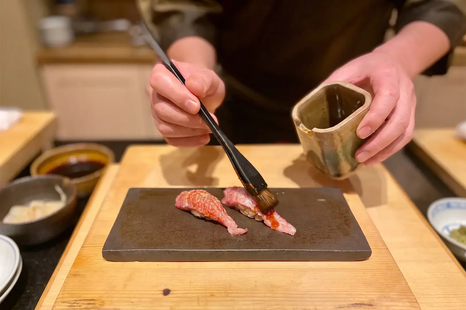 A chef carefully preparing a sushi meal with a brush and container on a wooden tray.