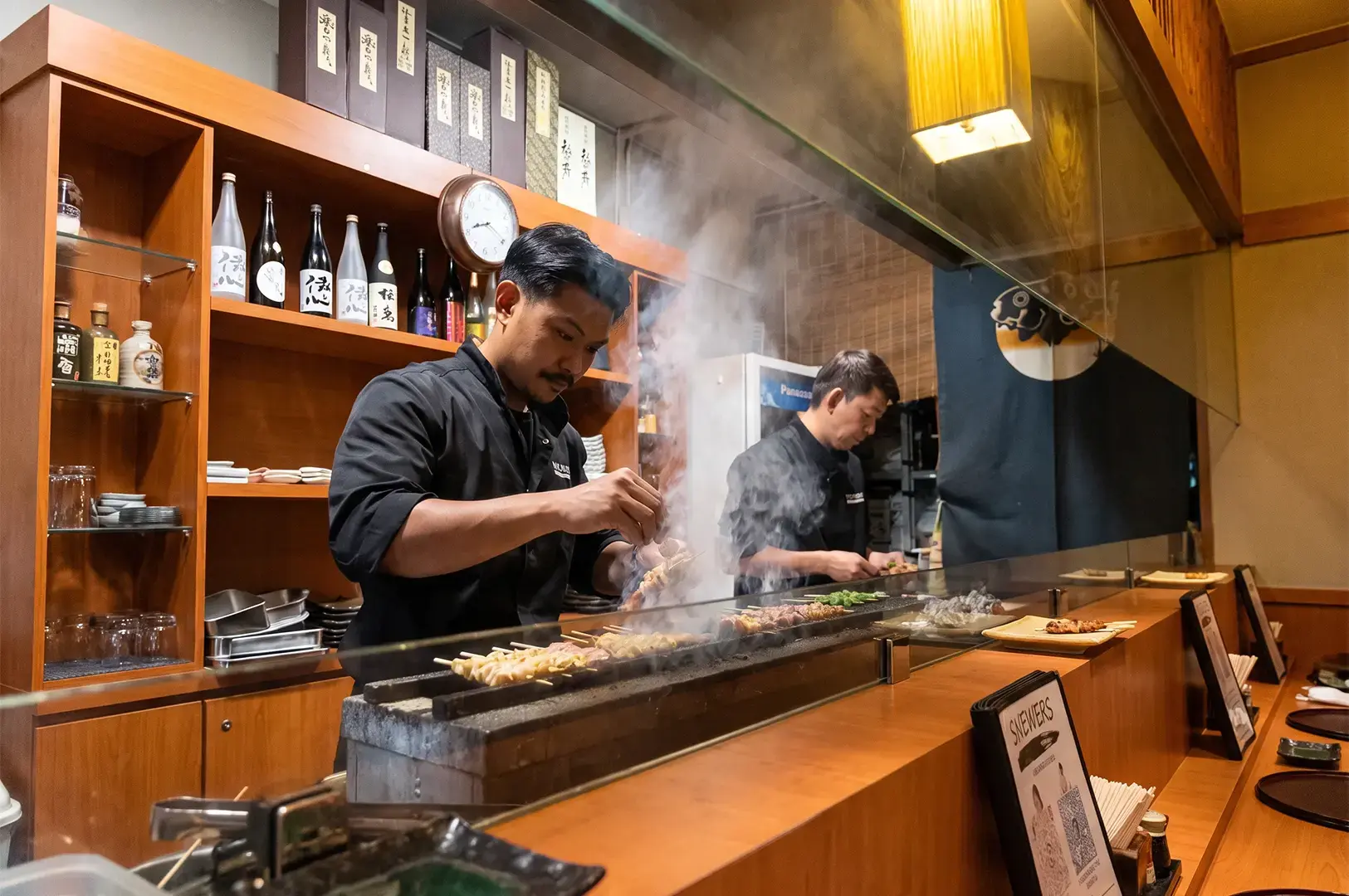 Two Skewers Yakitori staff grilling a large number of skewers, white smoke rising from the grills and platters and bottles arranged on the shelf behind.