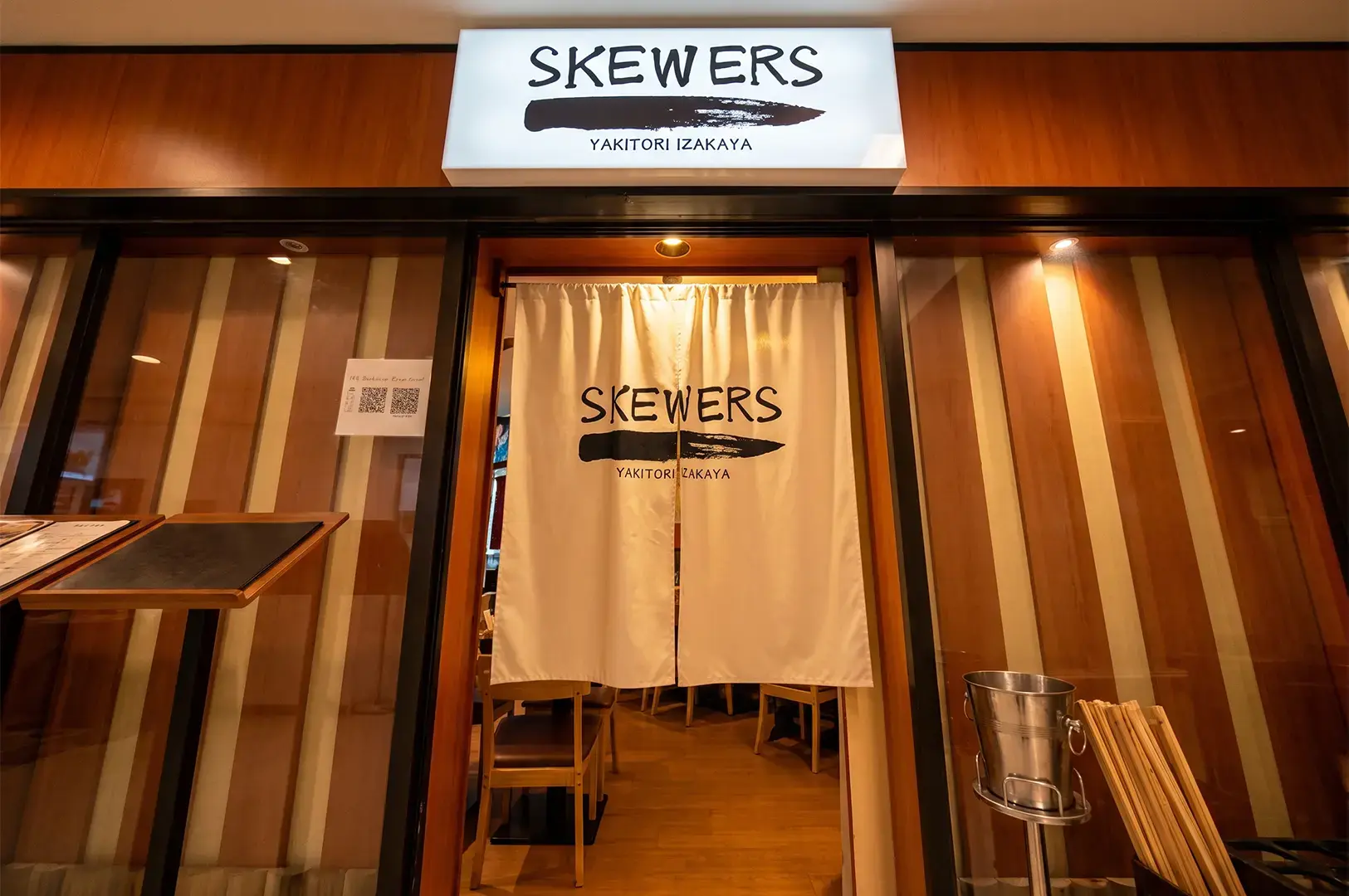 Wide angle shot of Skewers Yakitori Entrance with its upper signage and curtain decorated with their logo and wooden panels behind glass stretches out on either side.