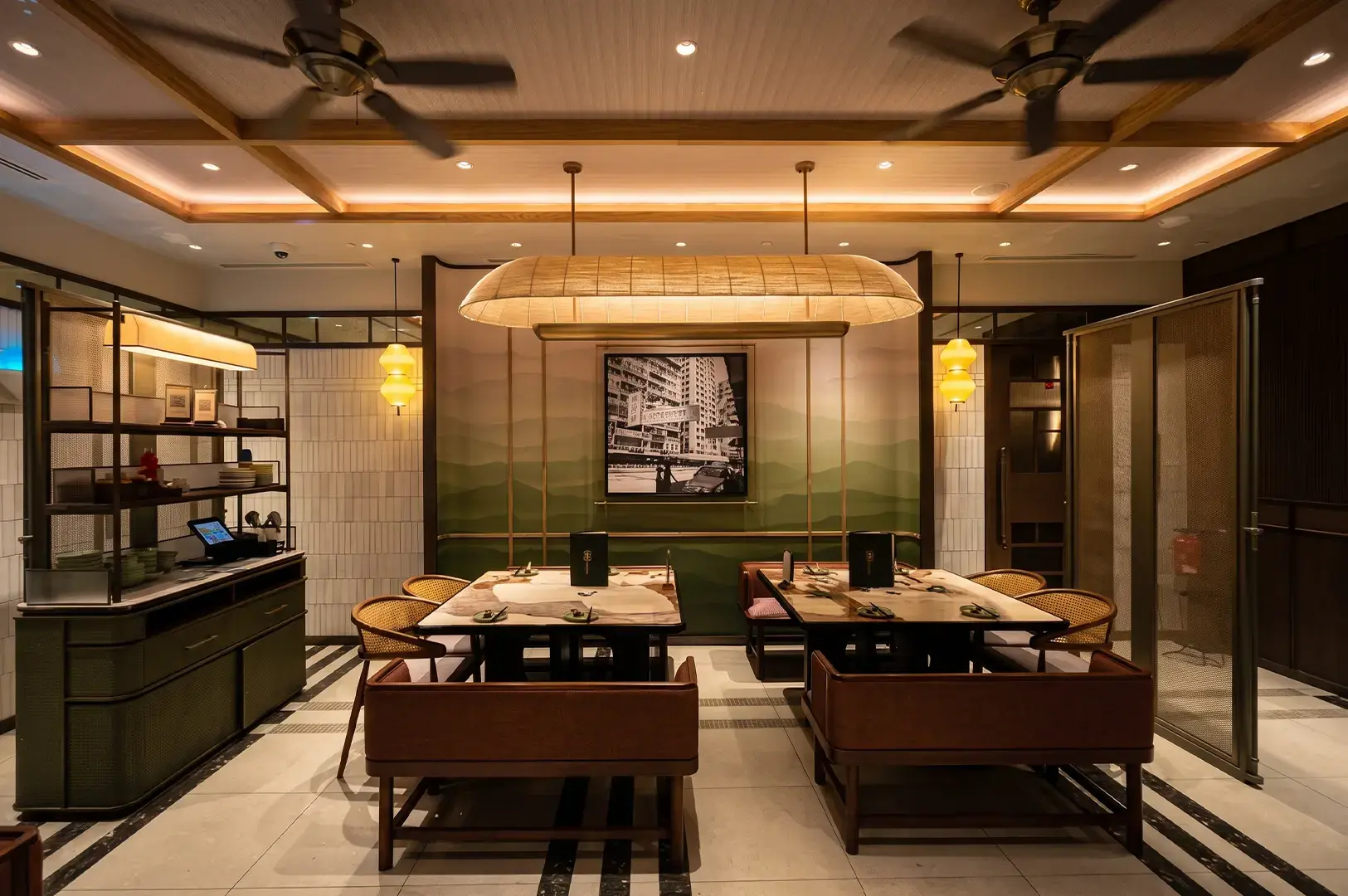 Wide angle shot of Tim Ho Wan interior with empty brown chairs and tables surrounded by panels with faint green paining textures illuminated by warm yellow lighting.