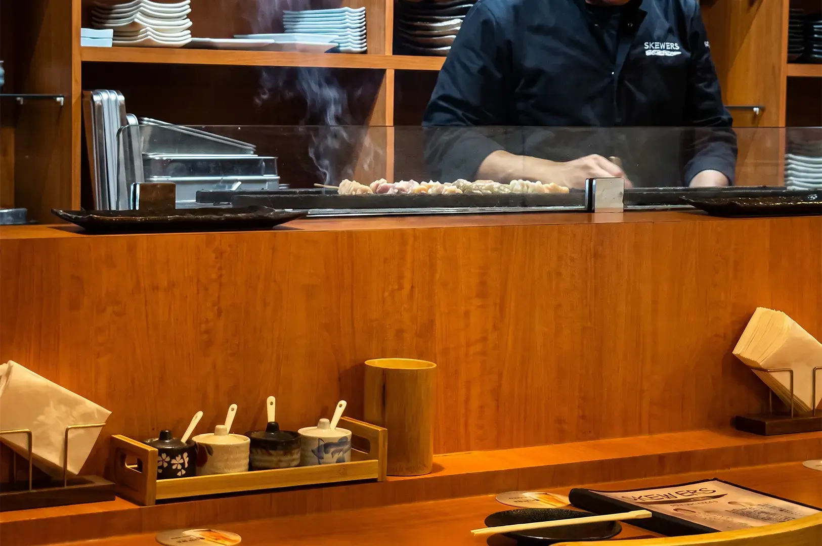 Close up shot of the grills inside Skewers Yakitori. with a staff grilling skewers and the different spices and utensils visible in the foreground.