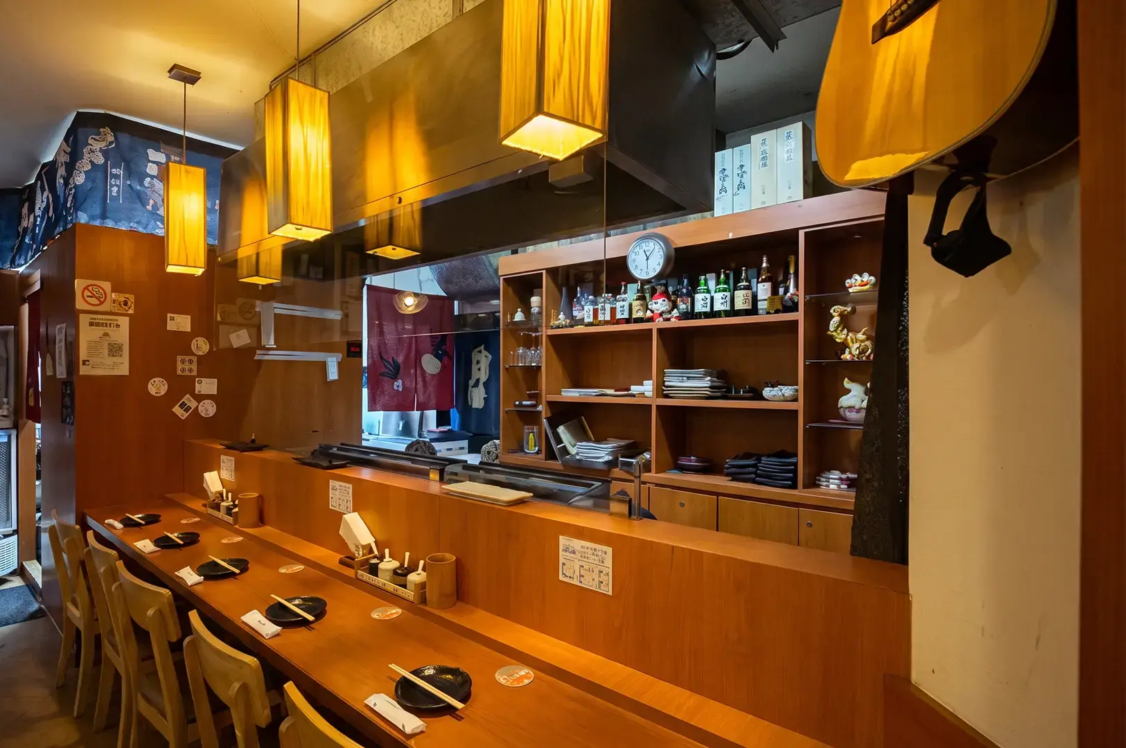 Skewers Yakitori interior with empy chairs and tables in front of the grills and the plates and bottles placed on shelves at the back, illuminated by warm yellow lights coming from rectangular hanging lanterns.