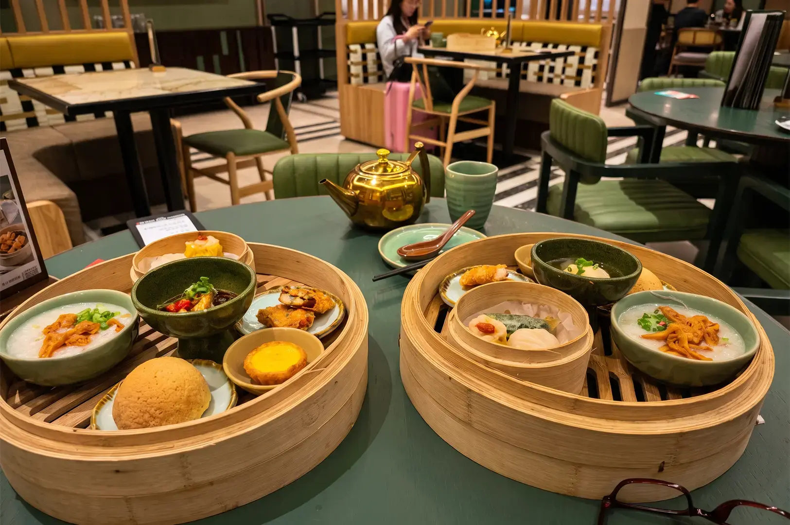 Tim Ho Wan grouped meals with pork buns, dim sum, and dumplings arranged in bamboo steamers on a dark green table, with other chairs and tables visible in the background.
