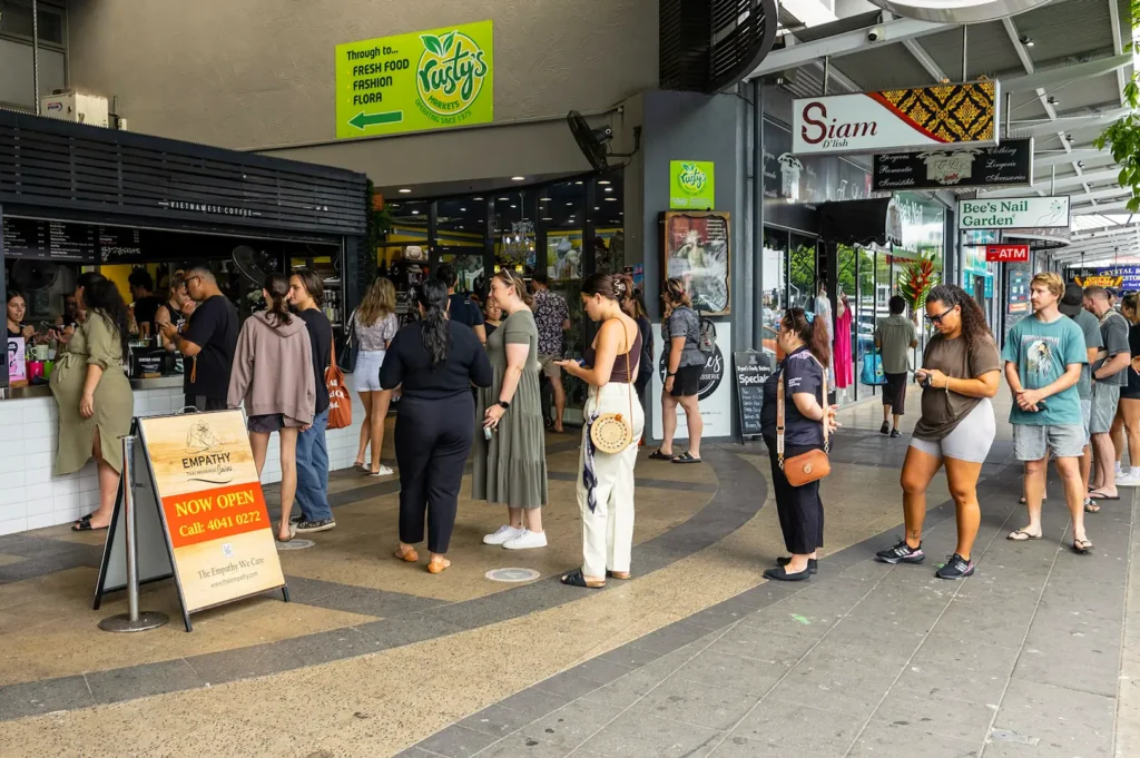 A line of customers in front of a cafe along a busy commercial street.